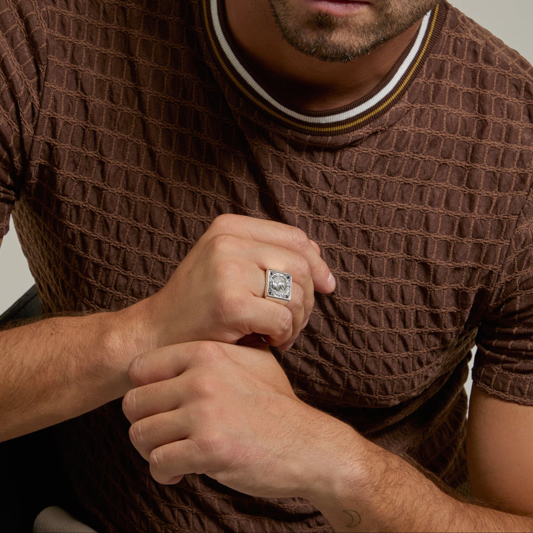 Man wearing a brown textured sweater and a ring, sitting against a neutral background