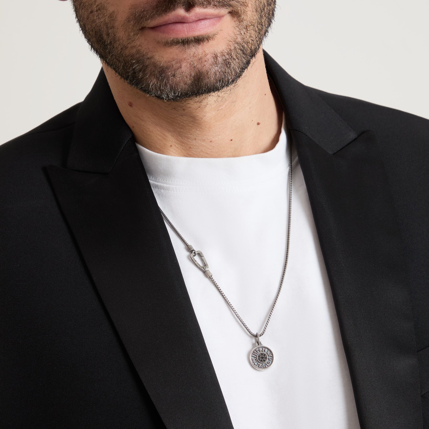 Man wearing a black blazer and white shirt with a silver necklace featuring a circular pendant.