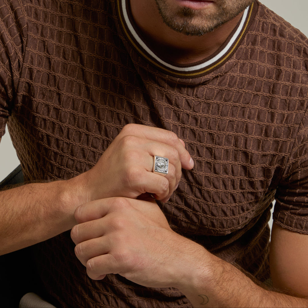 Man wearing a brown textured sweater and a ring, sitting against a neutral background