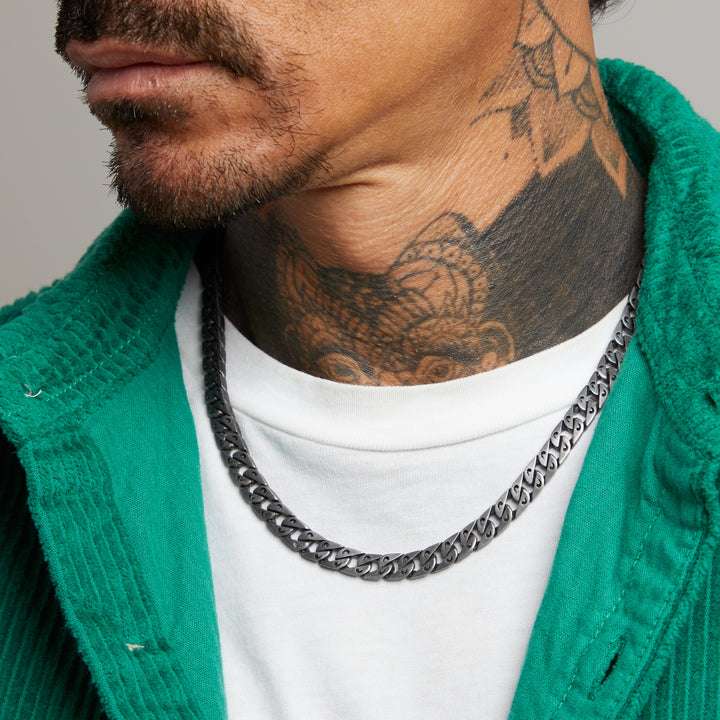Man wearing a green sweater and white shirt with a silver chain necklace, close-up on neck and tattoo.