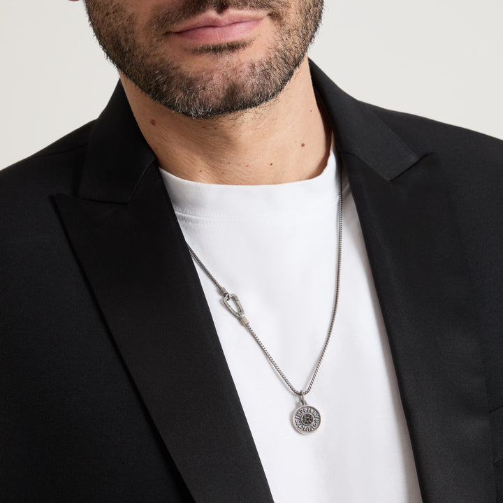 Man wearing a black blazer and white shirt with a silver necklace featuring a circular pendant.