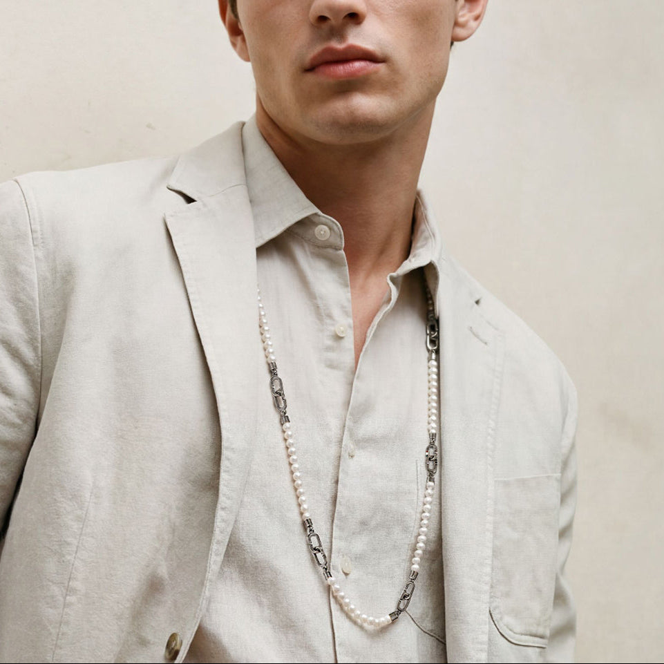 Man wearing a light-colored suit and shirt with a necklace against a plain background