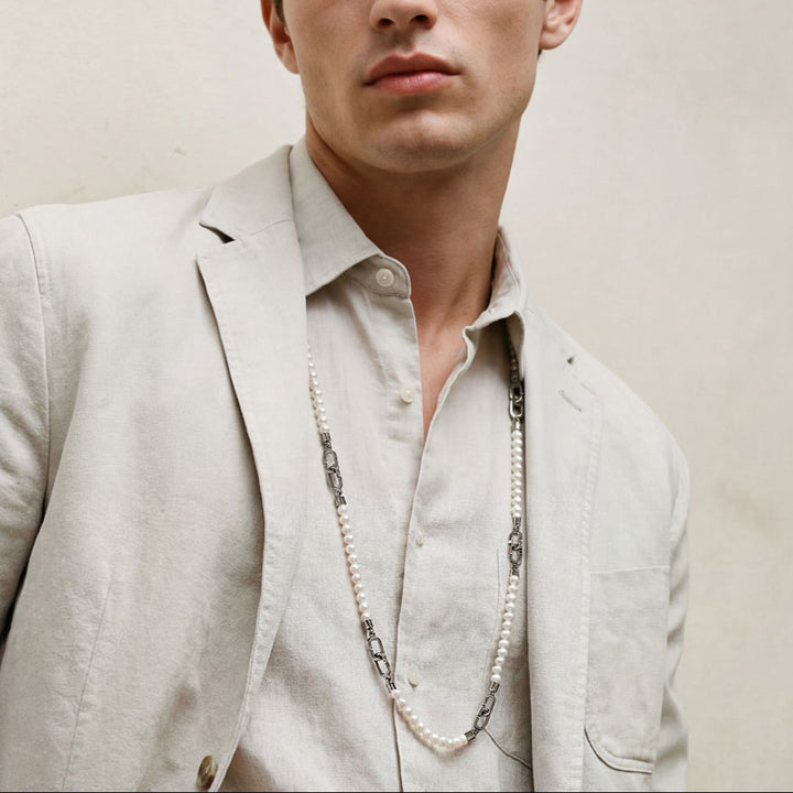 Man wearing a light-colored suit and shirt with a necklace against a plain background