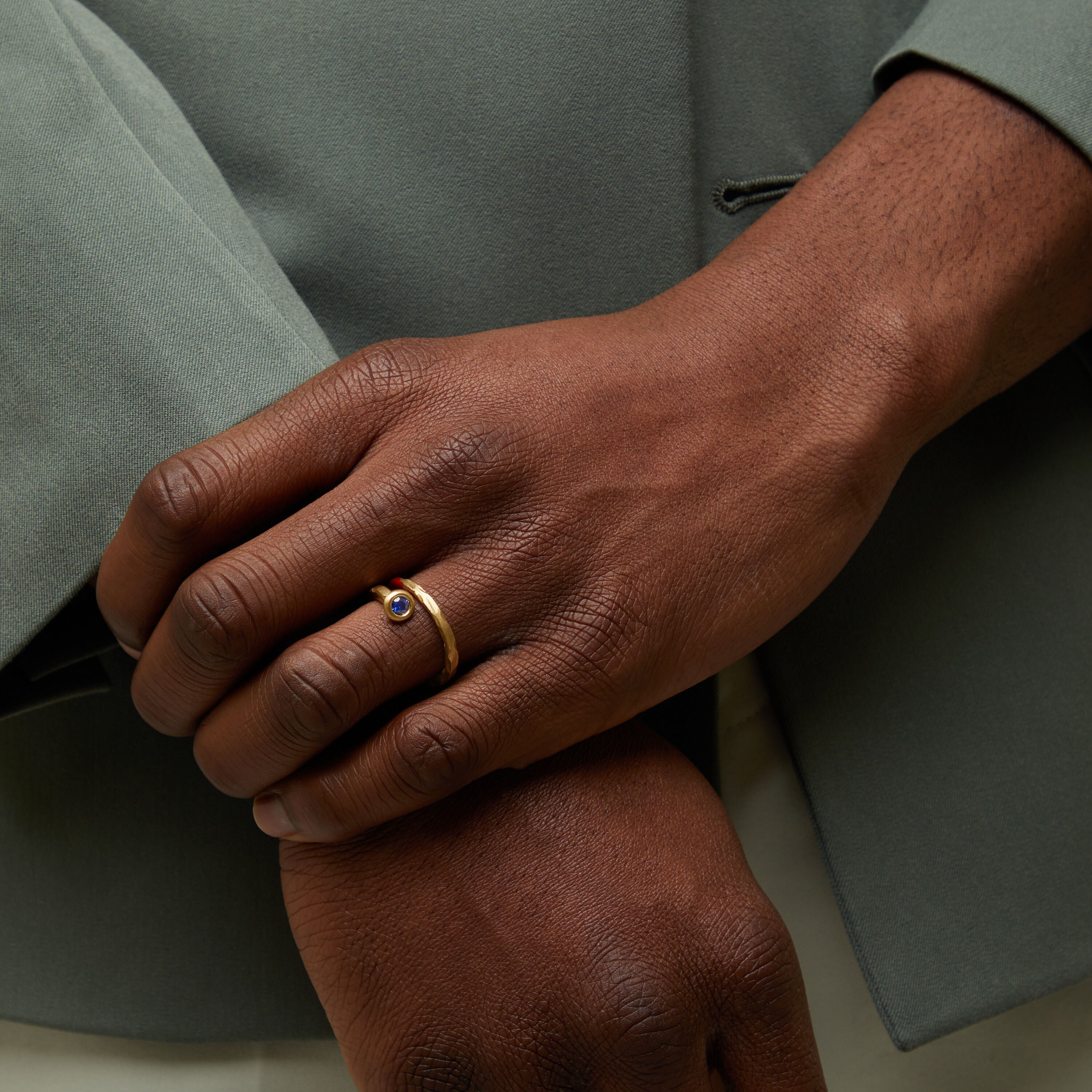 Close-up of a hand wearing a gold ring with a blue stone, against a neutral background.