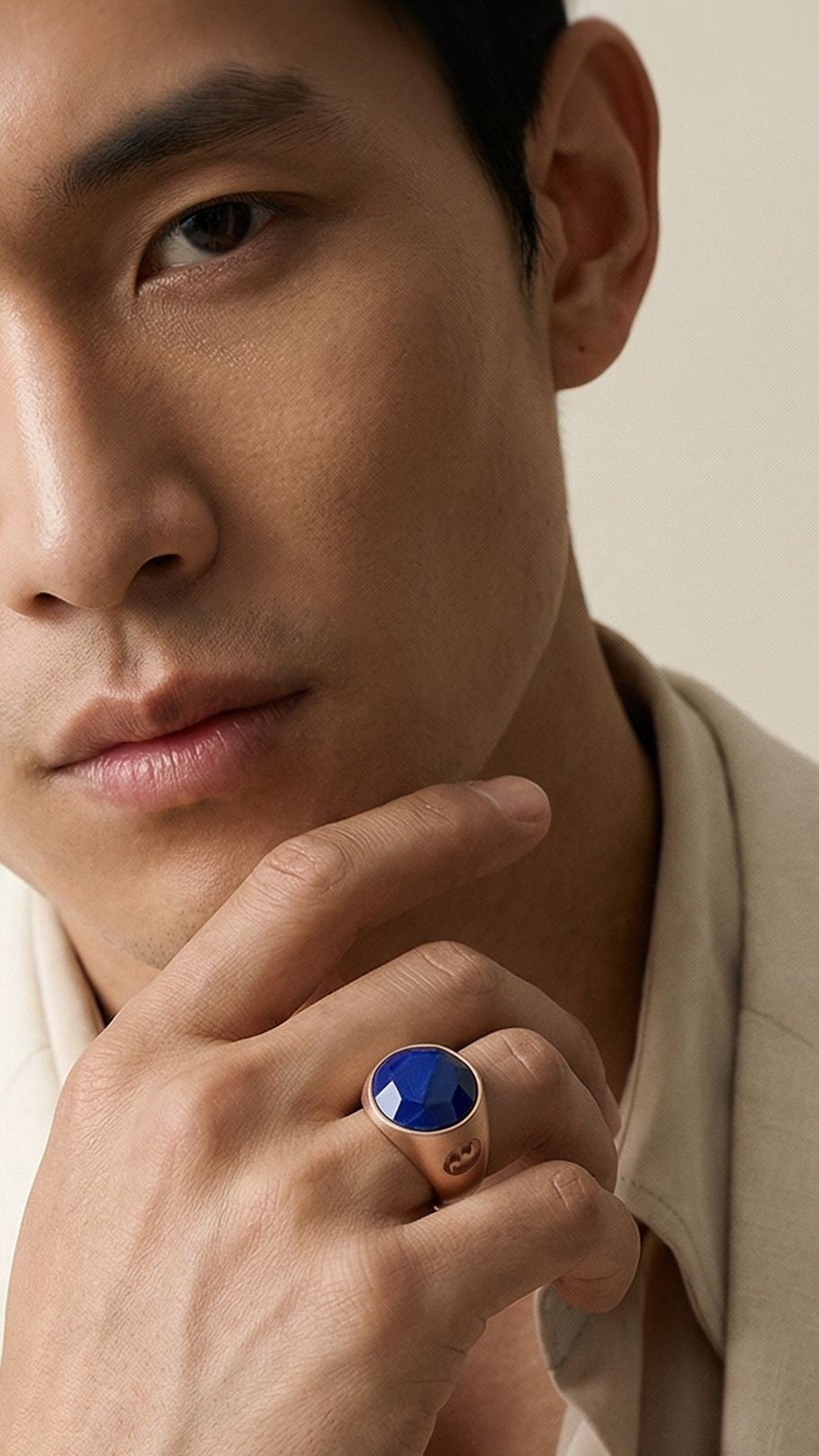 Man in a beige suit with a blue ring on his finger against a plain background