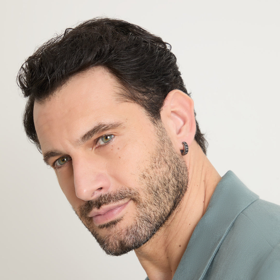 Male model wearing the MARCO DAL MASO Acies Mini Hoop Earring in oxidized silver with black enamel detailing, shown in a close-up portrait.
