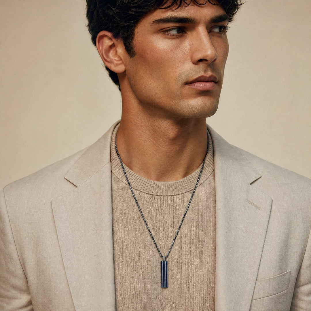 Male model wearing the MARCO DAL MASO Acies Pendant in oxidized sterling silver with blue enamel detailing, styled over a beige knit sweater and tailored jacket.