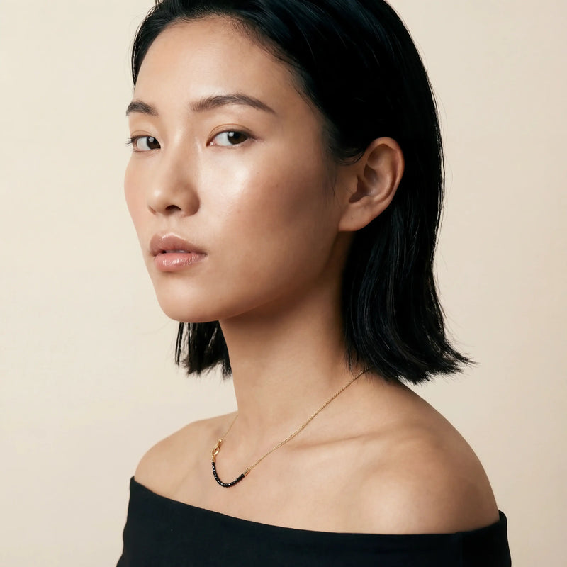 Female model wearing the MARCO DAL MASO XOK Wave Mini Beaded Chain in 18k yellow gold, featuring a delicate chain with a curved dark beaded detail, shown in a close-up portrait.