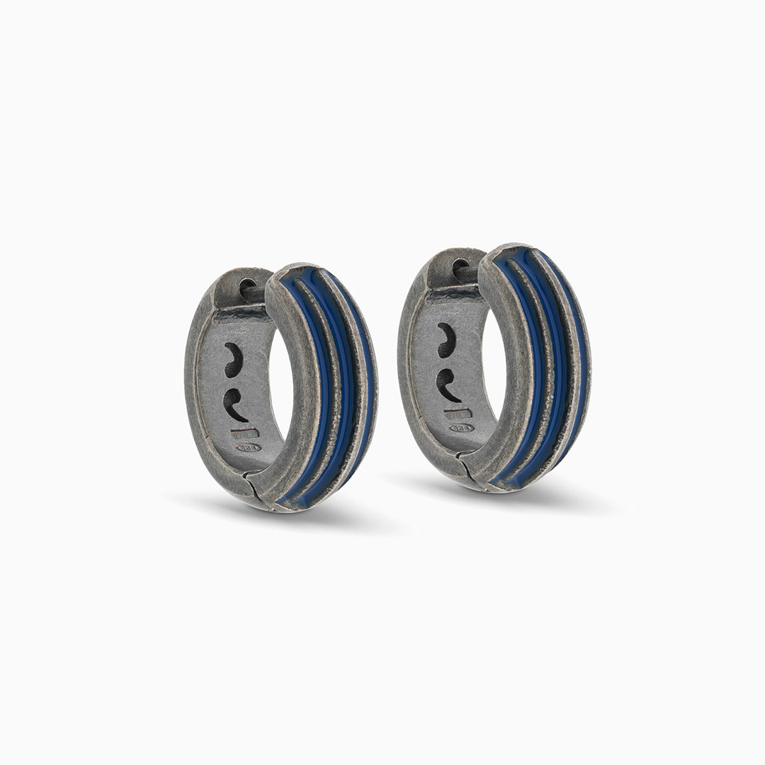 Pair of MARCO DAL MASO ACIES Huggie Earrings in oxidized silver with contrasting blue enamel stripes, featuring a curved hoop design against a light background.