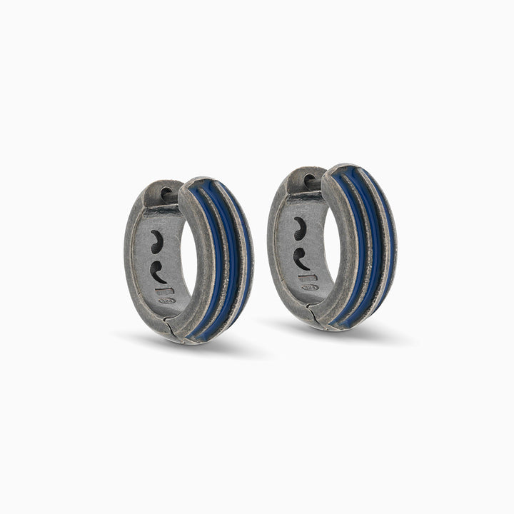 Pair of MARCO DAL MASO ACIES Huggie Earrings in oxidized silver with contrasting blue enamel stripes, featuring a curved hoop design against a light background.