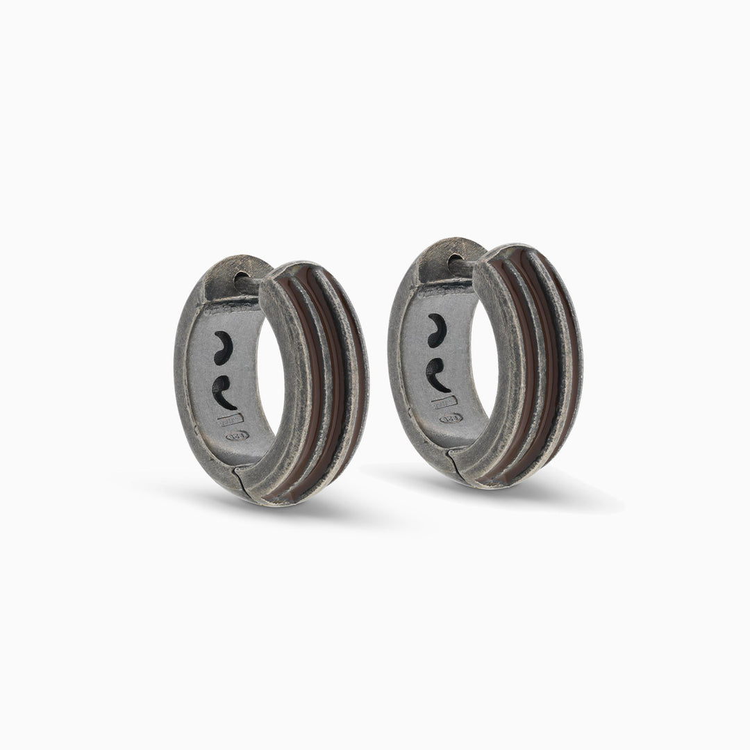 Pair of MARCO DAL MASO Acies Huggie Earrings in oxidized silver with brown enamel stripes, featuring a curved hoop design and textured metal finish, displayed against a light background.