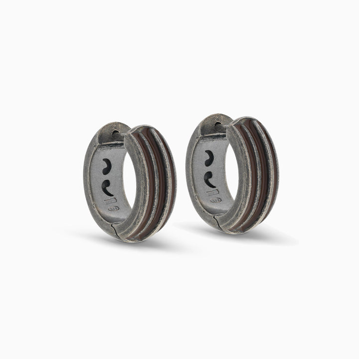 Pair of MARCO DAL MASO Acies Huggie Earrings in oxidized silver with brown enamel stripes, featuring a curved hoop design and textured metal finish, displayed against a light background.